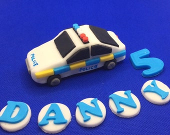 May include: A white police car cake topper with blue and yellow accents. The car has a black roof and windows. The words "Police" are written on the sides of the car. The cake topper is surrounded by white circles with blue letters spelling out the name "Danny" and a blue number "5".