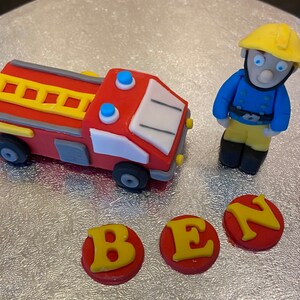May include: A red fire truck cake topper with a yellow ladder, blue lights, and white details. A yellow and blue firefighter figurine stands next to the truck. Three red and yellow fondant letters spell out "BEN".