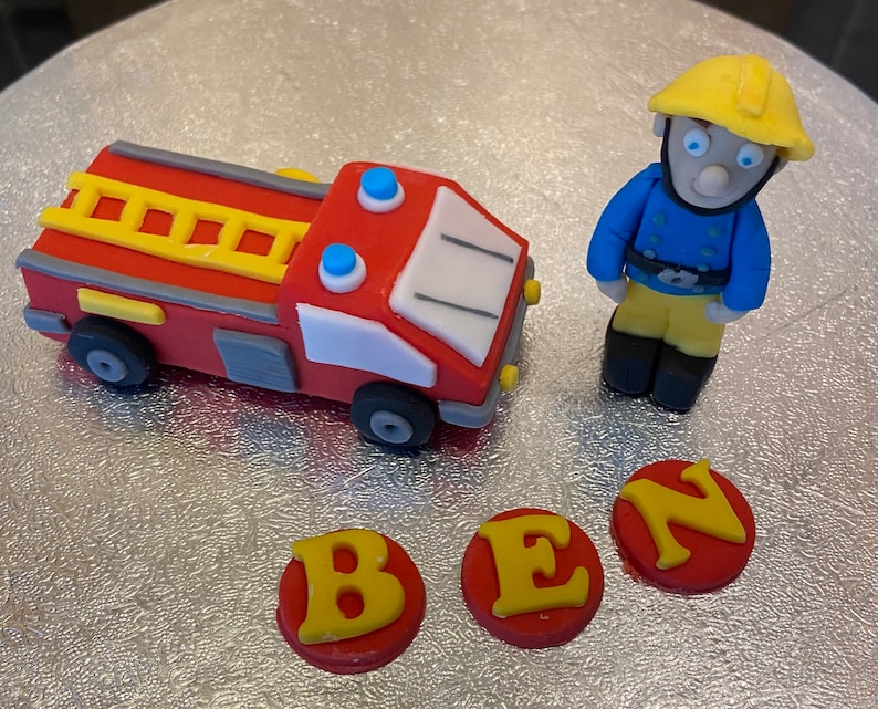 May include: A red fire truck cake topper with a yellow ladder, blue lights, and white details. A yellow and blue firefighter figurine stands next to the truck. Three red and yellow fondant letters spell out "BEN".