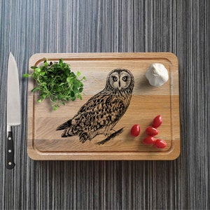 May include: A wooden rectangular chopping board with an engraved owl design. The board has a groove around the edge and is surrounded by fresh herbs, tomatoes, a garlic bulb, and a stainless steel knife.