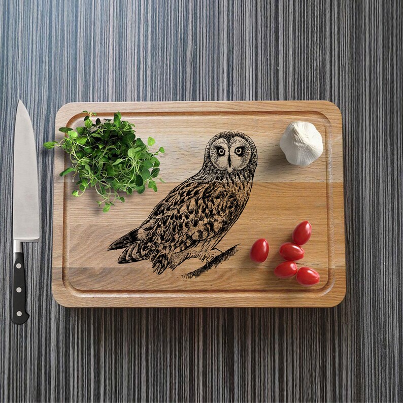 May include: A wooden rectangular chopping board with an engraved owl design. The board has a groove around the edge and is surrounded by fresh herbs, tomatoes, a garlic bulb, and a stainless steel knife.