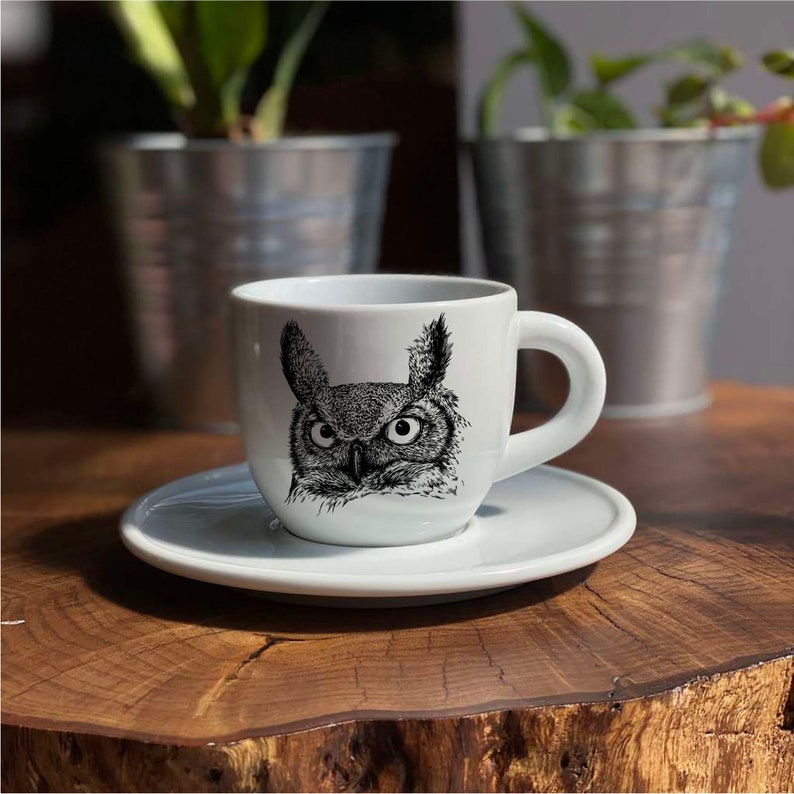May include: White ceramic coffee cup and saucer set. The cup features a detailed black and white illustration of an owl's face. The cup has a curved handle and sits on a matching saucer. The set is on a wooden surface.