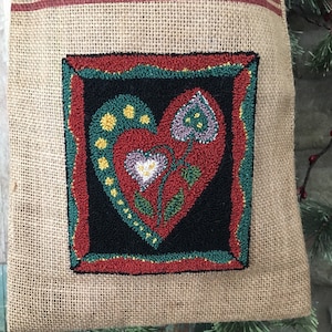 May include: A burlap stocking with a red and green embroidered heart design. The heart is surrounded by a red, green, and yellow frame.