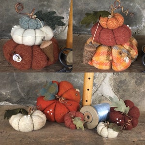 May include: A set of five handmade felt pincushions shaped like pumpkins. The pumpkins are in various sizes and colors, including orange, red, white, and blue. Some of the pumpkins have a plaid pattern. The pincushions are decorated with small felt leaves and acorns.