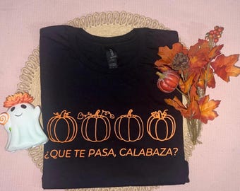 Puede incluir: Camiseta negra con diseño de calabazas naranjas y el texto "¿QUE TE PASA, CALABAZA?". La camiseta está doblada sobre una alfombra decorativa, con una figura de fantasma y hojas de otoño. Tema de Halloween.