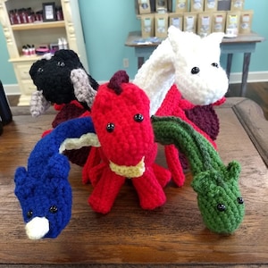 May include: Five crocheted stuffed dragons in different colors: red, blue, green, white, and black. The dragons are sitting on a wooden surface.