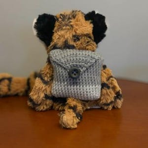 May include: A plush tiger toy with orange and black striped fur, black ears, and a long tail. The tiger is wearing a small, grey, crocheted square pouch on its chest, which features a single dark button closure at the centre of the flap.