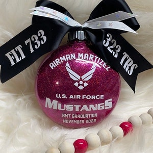 May include: Pink glitter Christmas ornament with the text "Airman Martizell" and "U.S. Air Force Mustangs BMT Graduation November 2022".  The ornament is decorated with a black ribbon with the text "FLT 733" and "323 TRS".