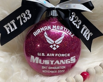 Op de afbeelding: Roze glitter kerstbal met de tekst "Airman Martizell" en "U.S. Air Force Mustangs BMT Graduation November 2022". De kerstbal is versierd met een zwart lint met de tekst "FLT 733" en "323 TRS".