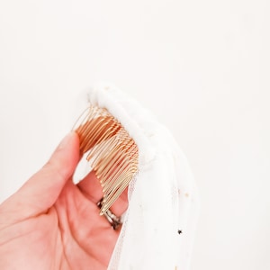 Op de afbeelding: Een gouden haar kam met een witte tule sluier eraan bevestigd. De sluier heeft gouden glittersterren.