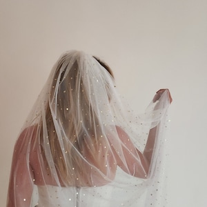 Op de afbeelding: Een witte tule sluier met gouden glittersterren die eroverheen zijn gestrooid. De sluier wordt gedragen door een persoon met lang blond haar.