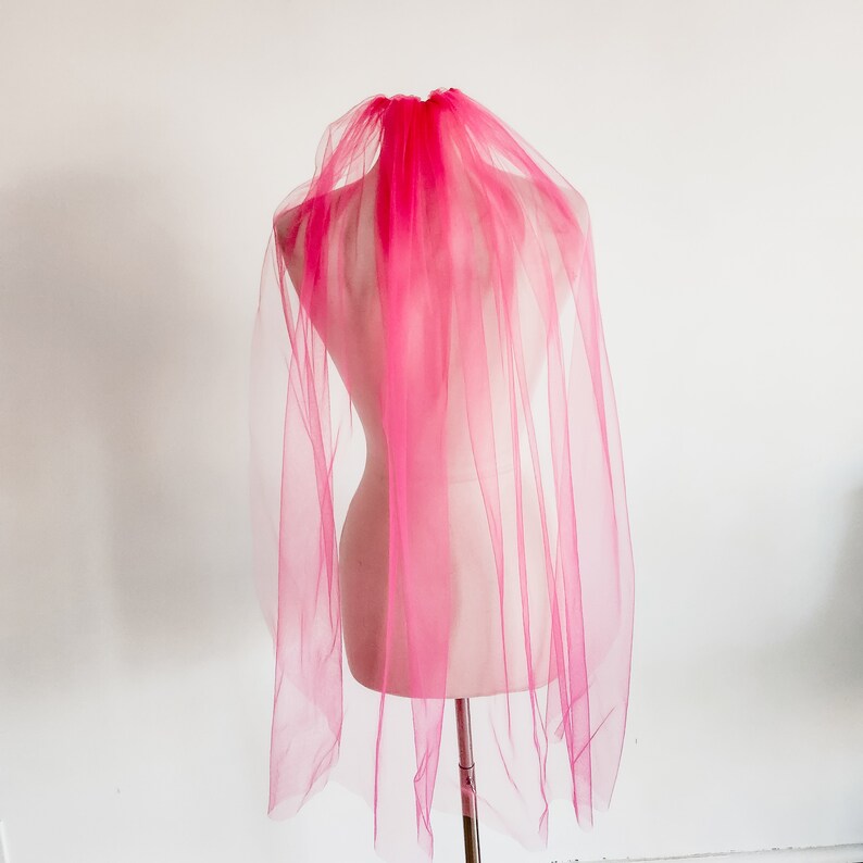 May include: A pink tulle veil with a single layer of fabric. The veil is draped over a mannequin's head and shoulders.