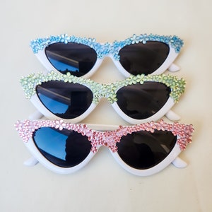 May include: Three pairs of white cat-eye sunglasses with different coloured flower embellishments. The sunglasses have black lenses.