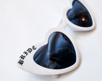May include: White heart-shaped sunglasses with black lenses and the word "BRIDE" printed on the frame.
