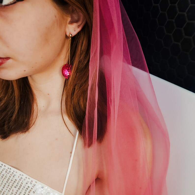 May include: A woman wearing a pink tulle veil and a white sequined top with a pair of dangling earrings featuring a pink sequin ball.