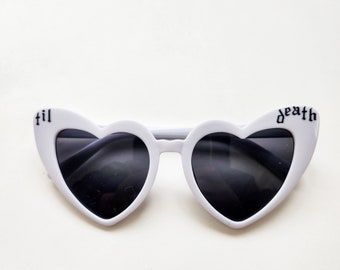 May include: White heart-shaped sunglasses with black lenses. The left lens has the word "til" printed on it and the right lens has the word "death" printed on it.