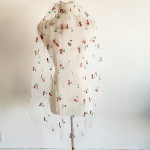 May include: A delicate white veil featuring embroidered red cherries and green leaves. The veil is displayed on a mannequin, highlighting its fine fabric and scattered fruit pattern. A bridal accessory.