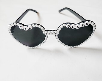 May include: Heart-shaped sunglasses with black lenses and a white beaded frame. The text "TIL DEATH" is spelled out in white letter beads across the top of the left lens, and "DEATH" is on the right lens. The frames are black.