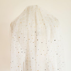 Op de afbeelding: Wit tule stof met gouden glittersterren. De stof is doorschijnend en heeft een delicate textuur.