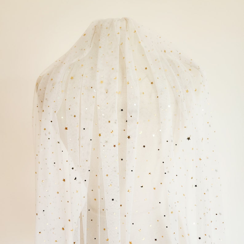 Op de afbeelding: Wit tule stof met gouden glittersterren. De stof is doorschijnend en heeft een delicate textuur.
