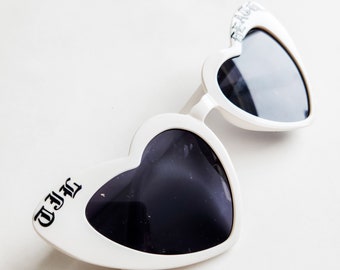 May include: White heart-shaped sunglasses with black lenses. The sunglasses have the text "T.J.H." on one side and "HE ART" on the other side.