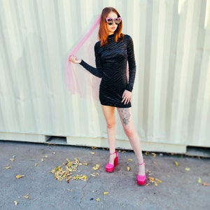 May include: A woman wearing a black velvet mini dress with a zebra print pattern. She is wearing pink heart-shaped sunglasses and a pink tulle veil. She is standing in front of a white wall with vertical lines. She is wearing pink high heels.