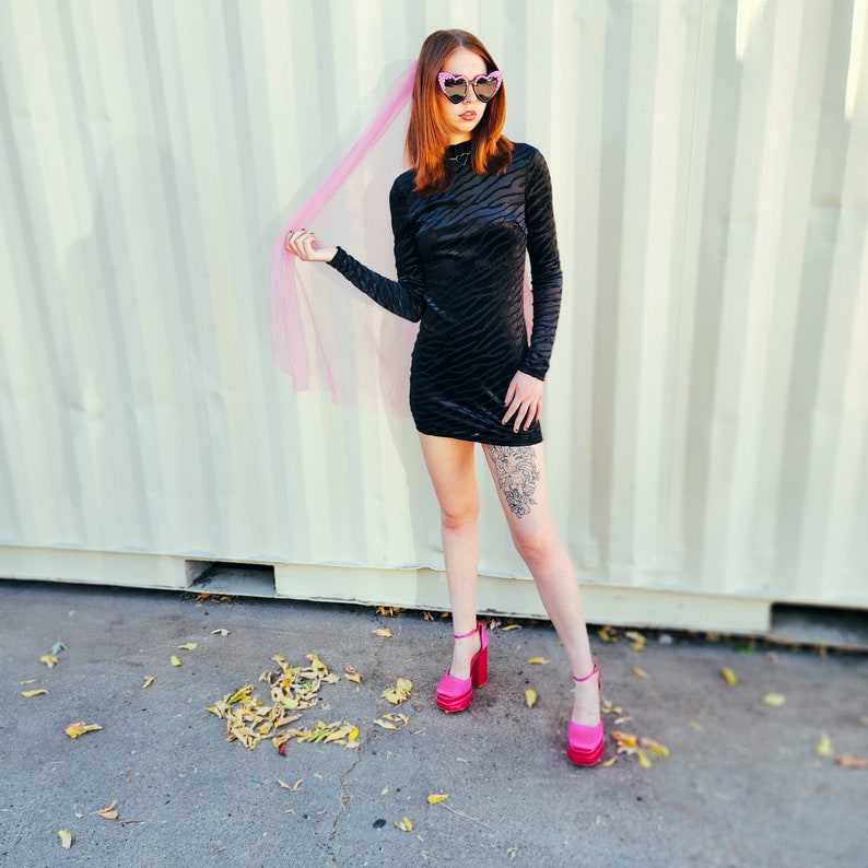 May include: A woman wearing a black velvet mini dress with a zebra print pattern. She is wearing pink heart-shaped sunglasses and a pink tulle veil. She is standing in front of a white wall with vertical lines. She is wearing pink high heels.