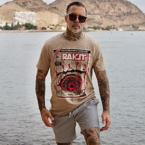 May include: A man wearing a tan t-shirt with a graphic print of a comic book cover that says "Synthesised Edition Rakit". He is wearing black and white striped shorts and has tattoos on his arms and legs.