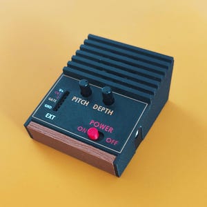 May include: A black electronic synthesiser module with a ribbed top section, featuring two black control knobs labelled PITCH and DEPTH. It includes a red power switch, a side input labelled EXT, and a wood-grain accent panel on the front edge.