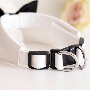 May include: White dog collar with a black bow tie and silver metal hardware. The collar is made of a soft, white fabric and has a buckle closure.