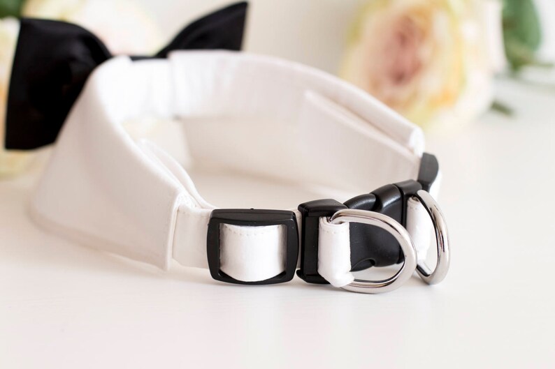 May include: White dog collar with a black bow tie and silver metal hardware. The collar is made of a soft, white fabric and has a buckle closure.