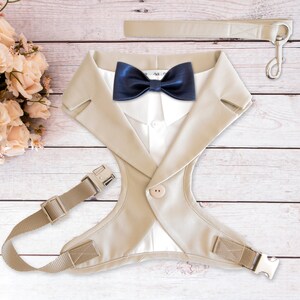 May include: A beige dog harness designed to resemble a suit jacket, complete with a navy blue bow tie. The harness has a white shirt-like front, a button closure, and adjustable straps. A matching beige leash with a silver clasp is also included.