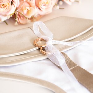 May include: A beige clutch with a white interior, adorned with two gold rings tied with a white ribbon. A bouquet of peach-colored roses rests in the background. The clutch has a beige strap.
