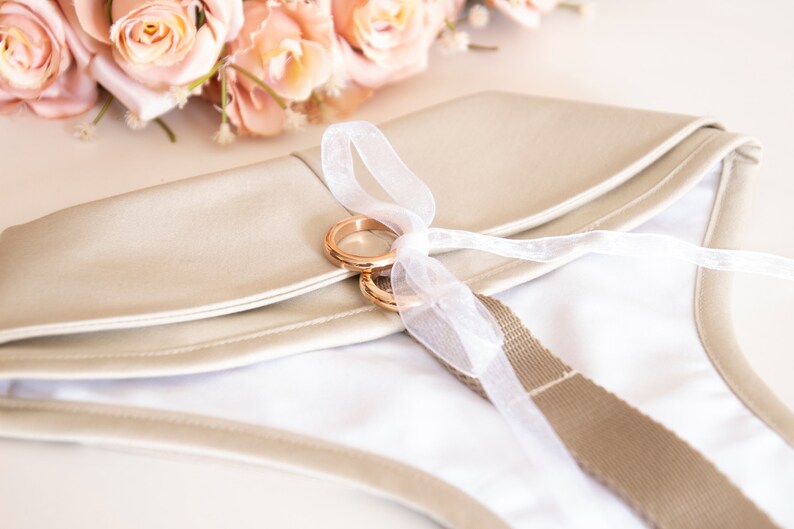May include: A beige clutch with a white interior, adorned with two gold rings tied with a white ribbon. A bouquet of peach-colored roses rests in the background. The clutch has a beige strap.
