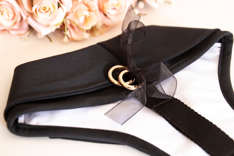 Puede incluir: Collar de tela negro con una cinta negra y dos anillos dorados. El collar est&aacute; unido a una correa negra.