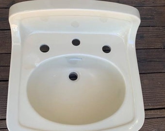 May include: A white ceramic bathroom sink with a single bowl and three holes for faucets. The sink has a curved backsplash and a rounded front edge.