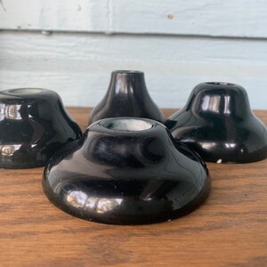 May include: Four glossy black candle holders with a rounded base and a shallow well for the candle. The candle holders are displayed on a wooden surface, with a light blue background.