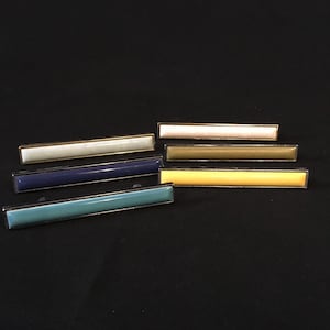 May include: Six different colored drawer pulls with a gold trim. The pulls are arranged in a row on a black surface. The colors are white, light green, dark blue, teal, yellow, and brown.