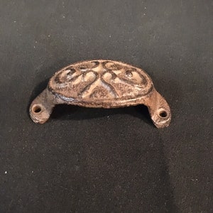 May include: A cast iron drawer pull with a decorative design. The pull is brown and has a slightly aged look.