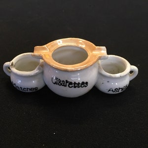 May include: A vintage white ceramic ashtray with three compartments. The center section is for cigarettes, with the word "Cigarettes" in black script. The side compartments are labeled "Matches" and "Ashes". The ashtray has a gold rim.