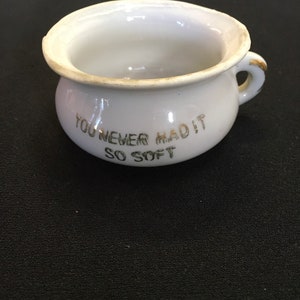 May include: A small, white ceramic chamber pot with a handle and the text "YOU NEVER HAD IT SO SOFT" in gold lettering. The rim and handle have a gold trim. The pot is on a black surface.