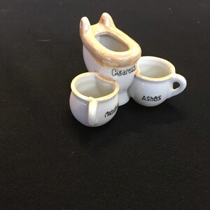 May include: A white ceramic ashtray set with a toilet-shaped cigarette holder and two small cup-shaped containers. The cigarette holder is labeled "Cigarette", one cup is labeled "Ashes", and the other "Matches".