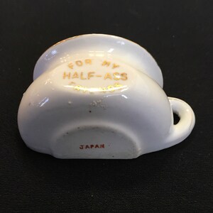 May include: A small, white ceramic ashtray with a handle and a curved rim. Gold lettering on the side reads "FOR MY HALF-ASS SAVINGS." The word "JAPAN" is printed in red on the bottom.