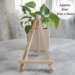 May include: A small wooden easel, approximately 9cm by 16cm, with a white pot and a green plant on top. The easel is made of natural wood and has a simple design. The text "@rainscraftybits" is visible at the bottom of the image.