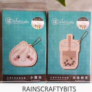 Puede incluir: Dos paquetes de kits de punto de cruz contados para hacer adornos de cuero. Los kits son para hacer un pequeño pan al vapor y un té de burbujas. Los paquetes son ambos azul claro con texto blanco y una pequeña imagen del producto terminado.