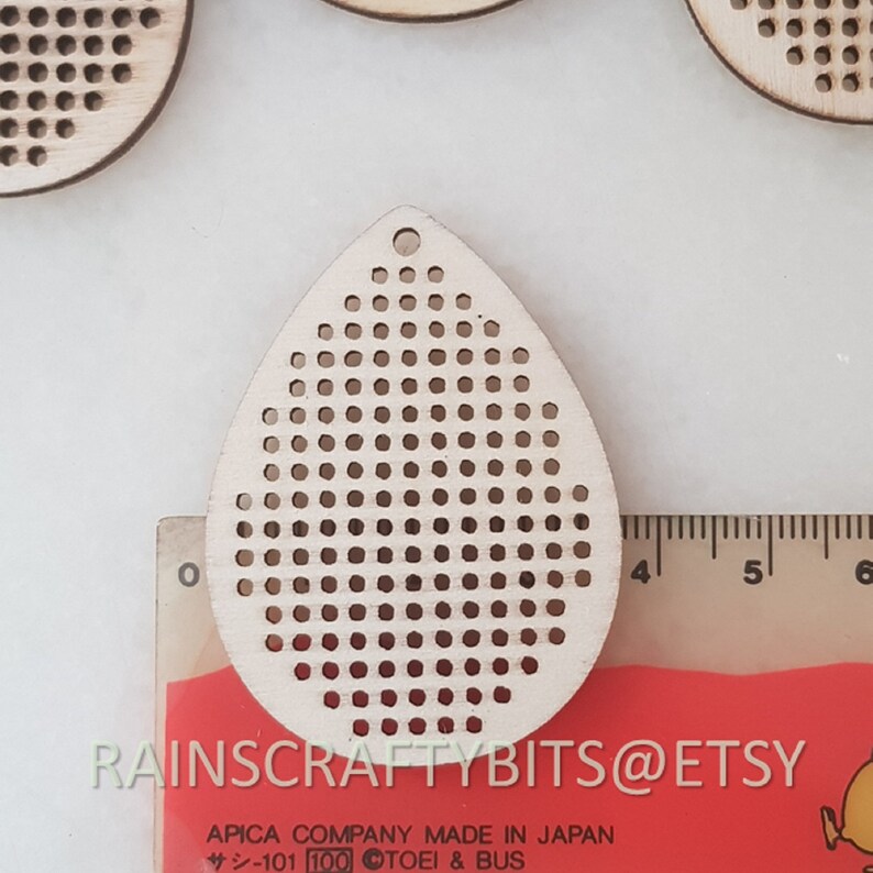 May include: A wooden teardrop-shaped craft pendant with numerous small, evenly spaced holes. The pendant has a small hole at the top for hanging. A ruler with inch measurements is visible. The text "RAINSCRAFTYBITS@ETSY" is also visible.