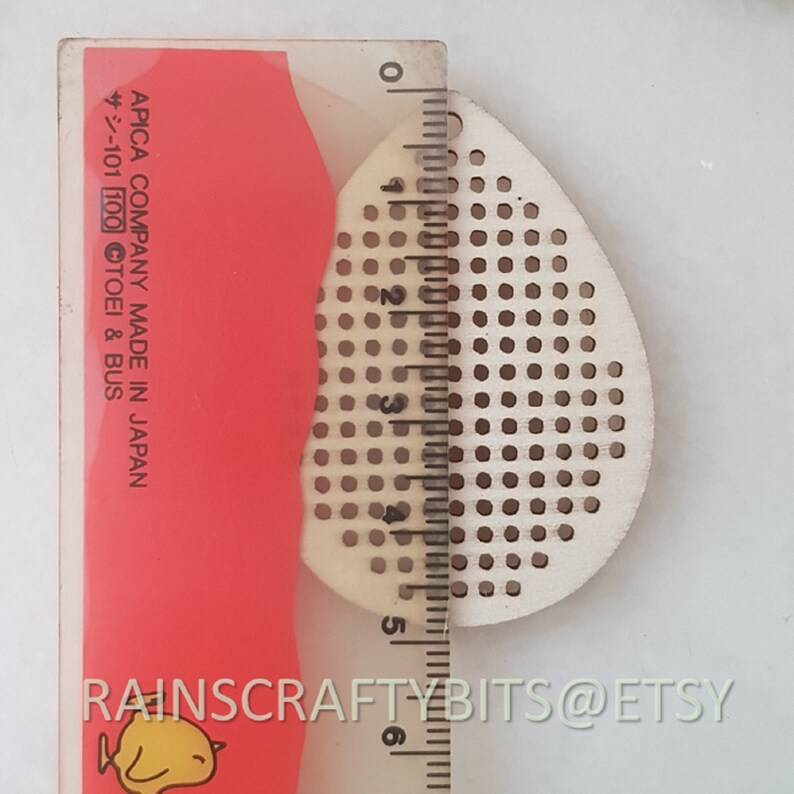 May include: A light-colored, teardrop-shaped wooden craft piece with evenly spaced holes, next to a ruler. The ruler shows measurements in inches. The text "RAINSCRAFTYBITS@ETSY" is visible at the bottom.
