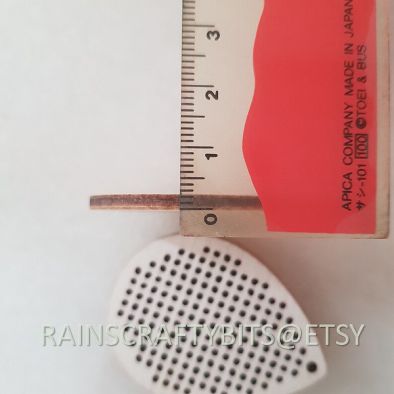 May include: A wooden teardrop-shaped craft supply with a perforated surface, next to a ruler showing a thickness of approximately 0.08 inches. The text "RAINSCRAFTYBITS@ETSY" is visible. A red and white ruler is also present.