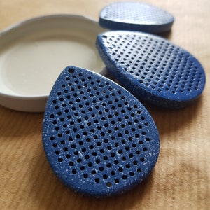 May include: Three blue teardrop-shaped objects with numerous small holes, speckled with white, are arranged near a white, oval-shaped dish. The objects have a textured appearance. The background is a light brown surface.