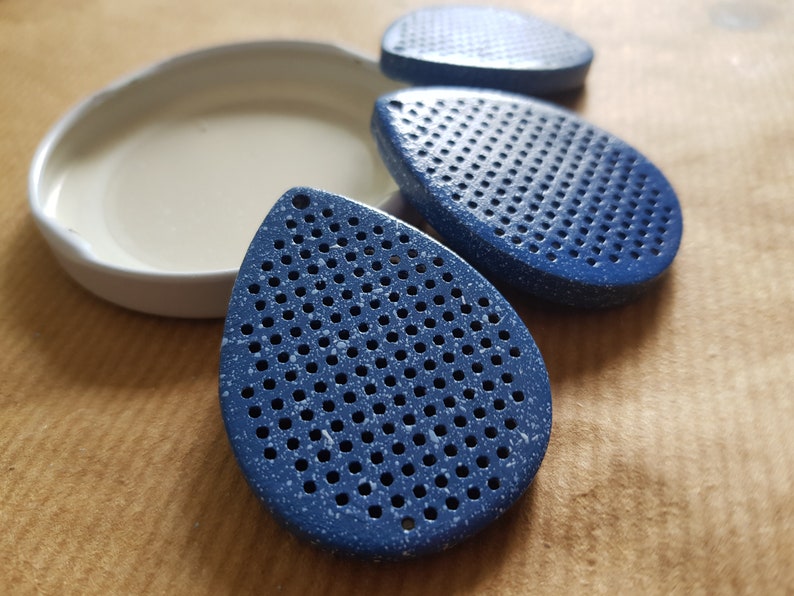 May include: Three blue teardrop-shaped objects with numerous small holes, speckled with white, are arranged near a white, oval-shaped dish. The objects have a textured appearance. The background is a light brown surface.